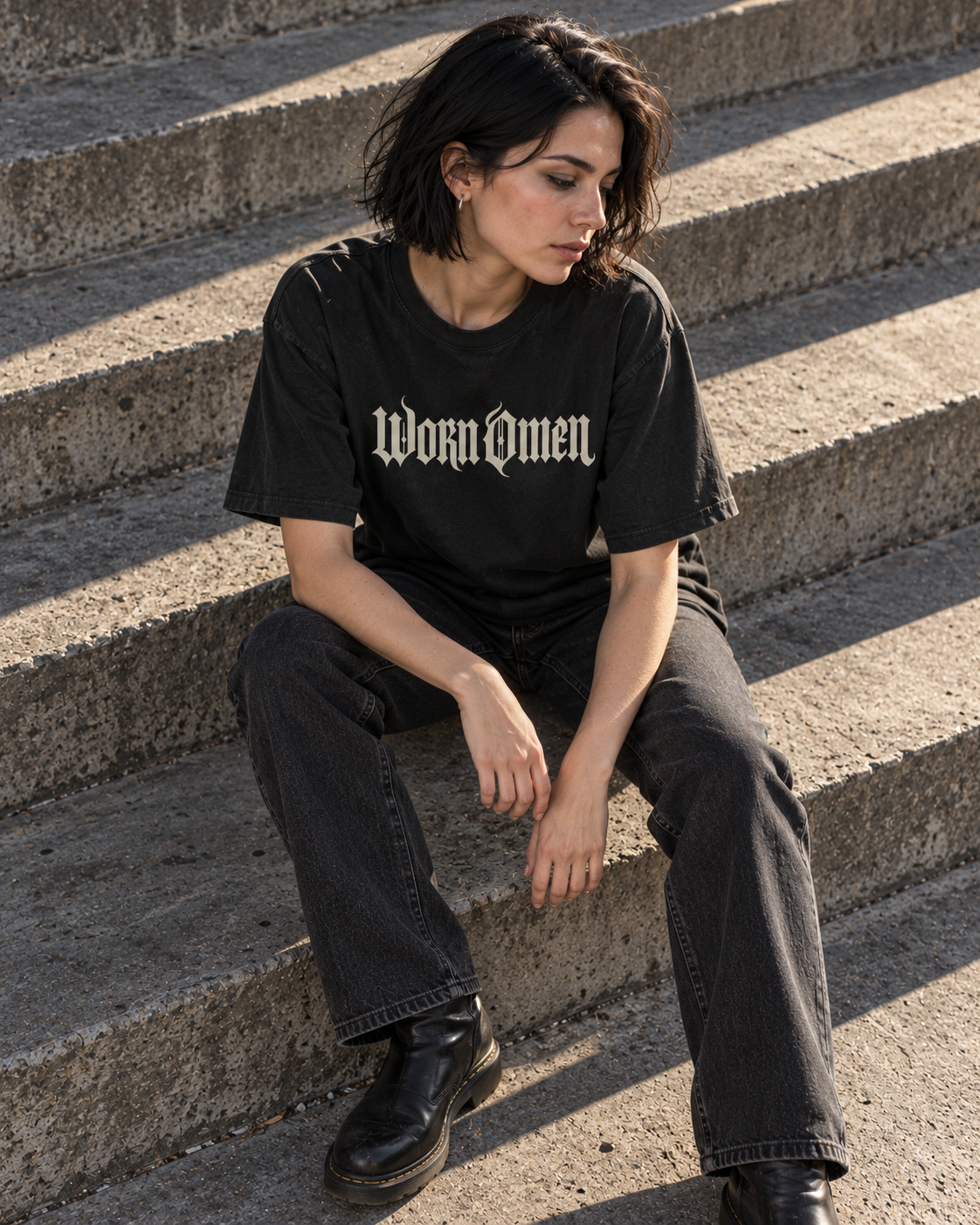 Seated model wearing the faded black WORN OMEN Signature Tee on concrete steps
