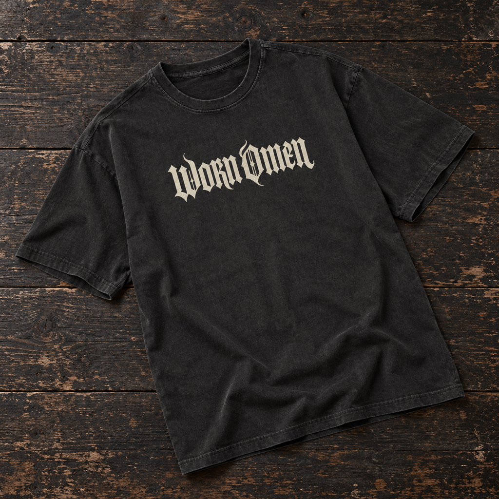WORN OMEN oversized gothic T-shirt in acid-washed black with bone lettering, laid on dark timber