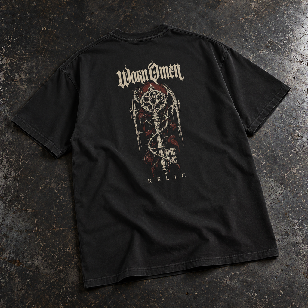 WORN OMEN Relic solid-black oversized tee with ornate key artwork on the back, laid on weathered steel